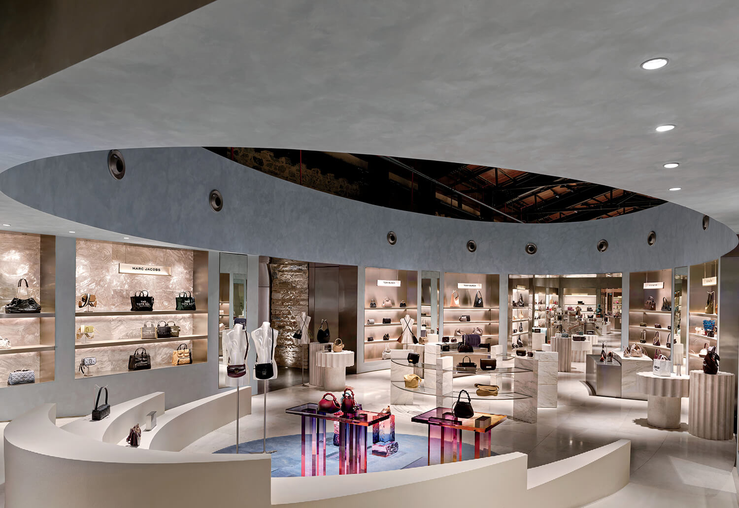 Luxury handbag and accessory store interior with curved display shelves, modern lighting, and various bags and items arranged on tables and stands.
