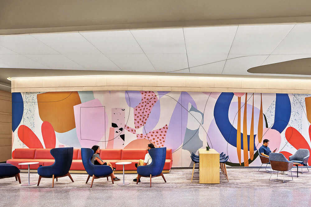 Modern lobby with colorful abstract mural, lounge chairs, red sofas, and people sitting; a wooden lectern stands near the center.