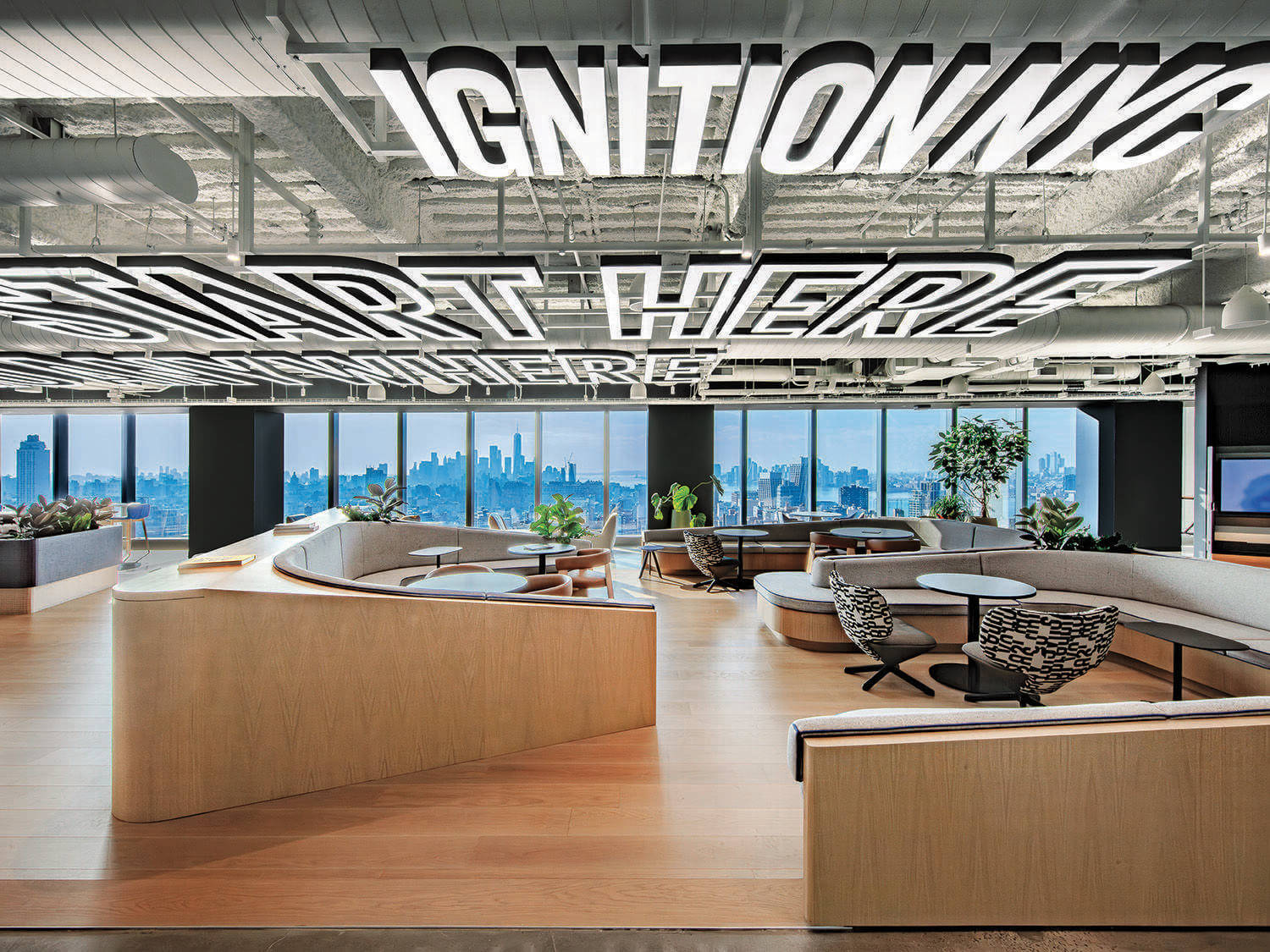 Modern office lounge with curved wooden seating, round tables, and large windows showing a city skyline. The ceiling displays bold white text reading “IGNITION NYC” and “START HERE.”.