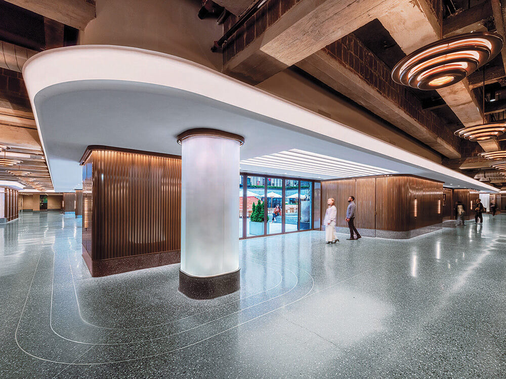 Two people walk through a modern lobby with polished floors, wood-paneled walls, a large column, and stylish lighting fixtures. Glass doors are visible in the background.