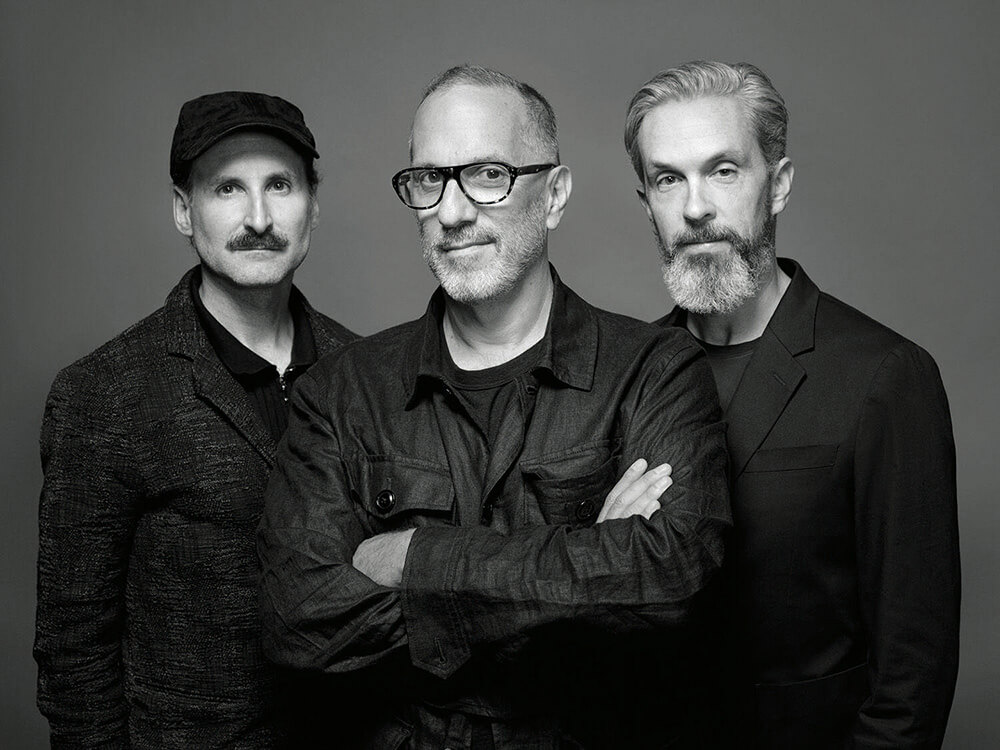 Three men with facial hair pose together in dark clothing against a plain background; the man in the center stands with arms crossed and wears glasses.