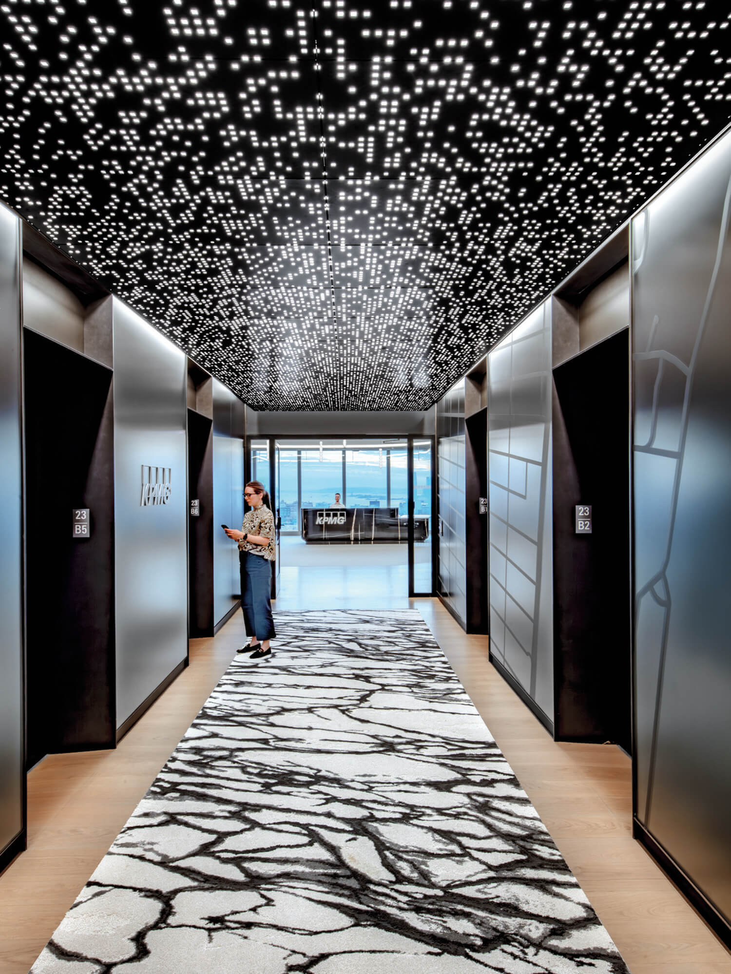 A person stands in a modern elevator lobby with a patterned rug, metallic walls, and a ceiling with a grid of small lights.