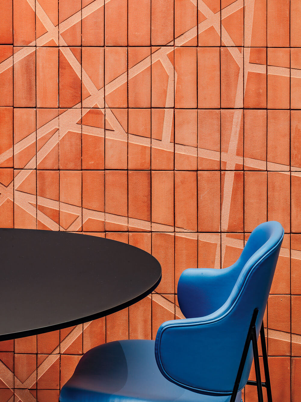 Blue chair and round black table in front of an orange tiled wall with intersecting line patterns.