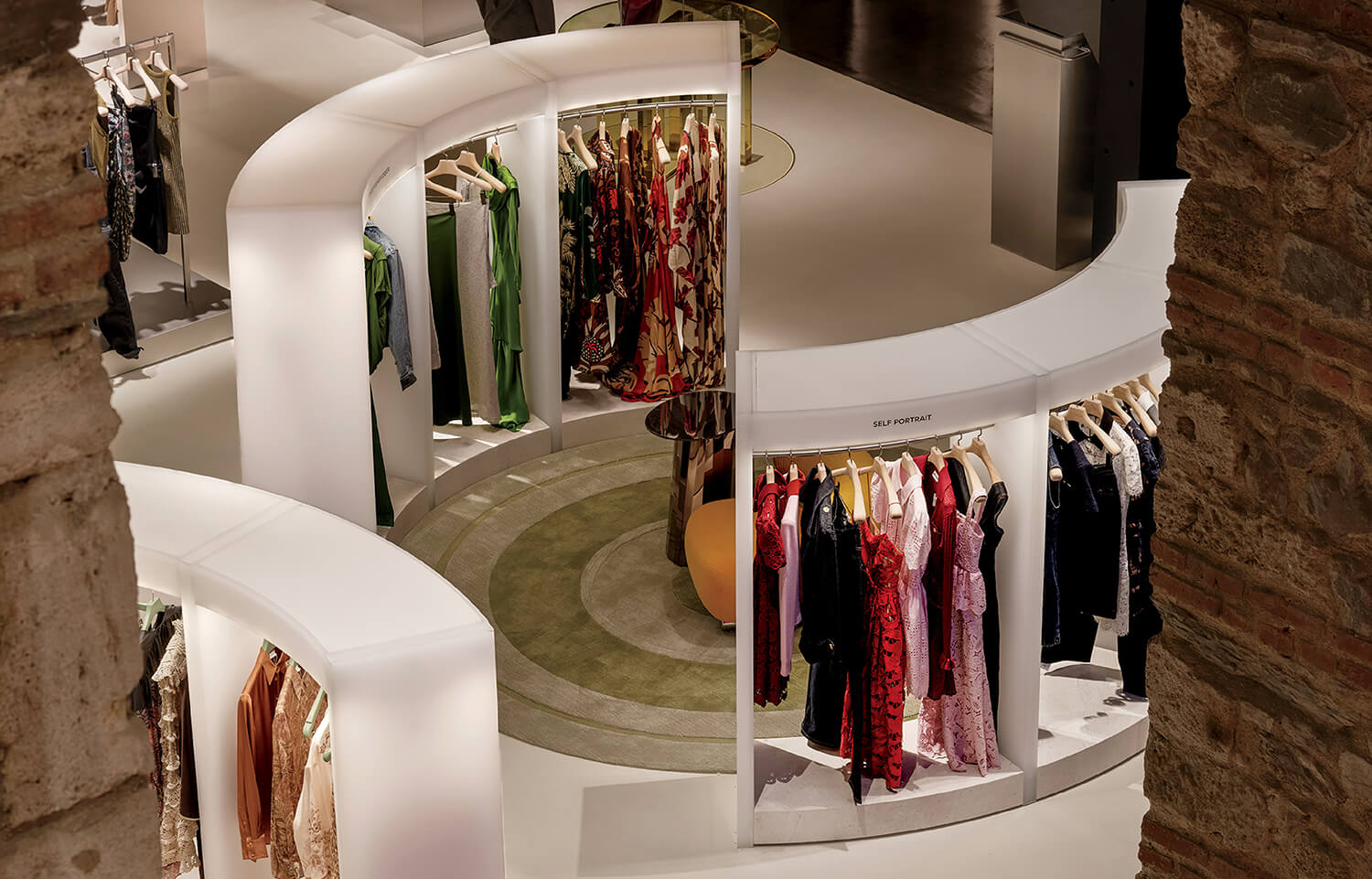 Curved white displays showcase assorted dresses on hangers in a modern clothing store, with stone walls partly visible in the foreground.