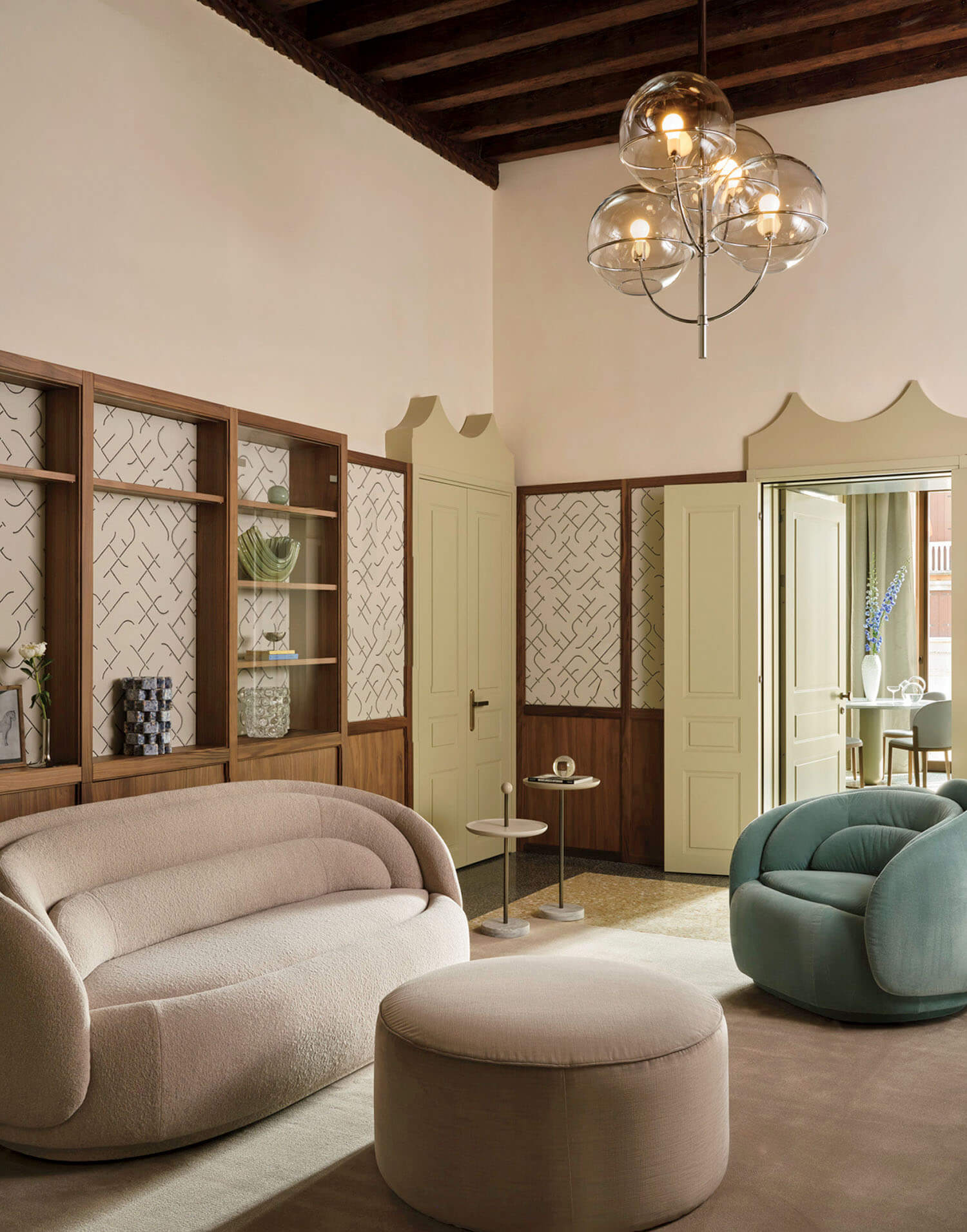 Modern living room with a curved beige sofa, round ottoman, teal armchair, wood shelving, and a glass bubble chandelier under a wooden ceiling.