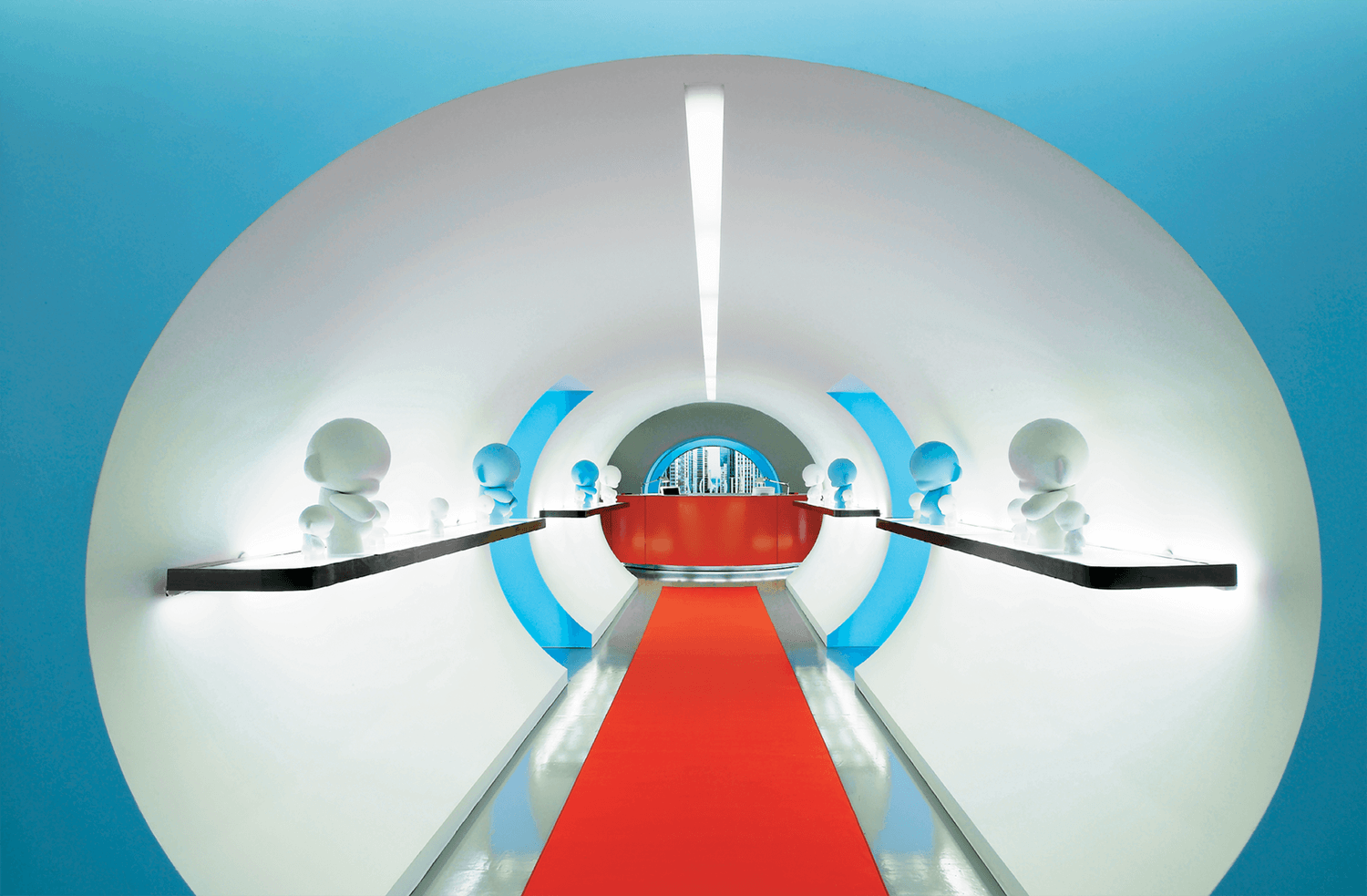 A futuristic white tunnel with a red carpet, lined with small humanoid statues on black shelves, leads to a window overlooking a cityscape.