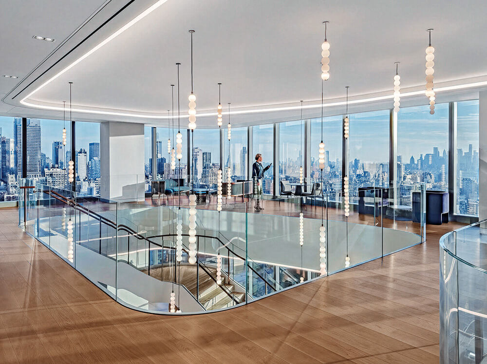 Modern office space with floor-to-ceiling windows, city skyline views, glass railings, pendant lights, and a person standing near the windows on the upper floor.