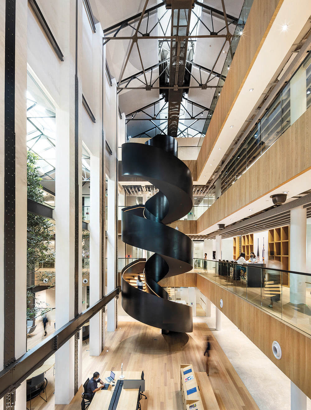 Modern atrium with a central black spiral staircase, high ceilings, glass roof, wood floors, and people working at desks and walking.