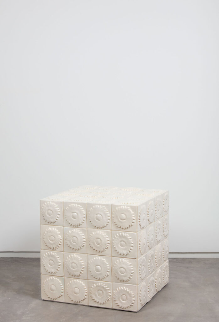 White cube stool with a grid pattern, each square featuring a raised circular, sunburst-like design, placed on a gray concrete floor against a plain white wall.