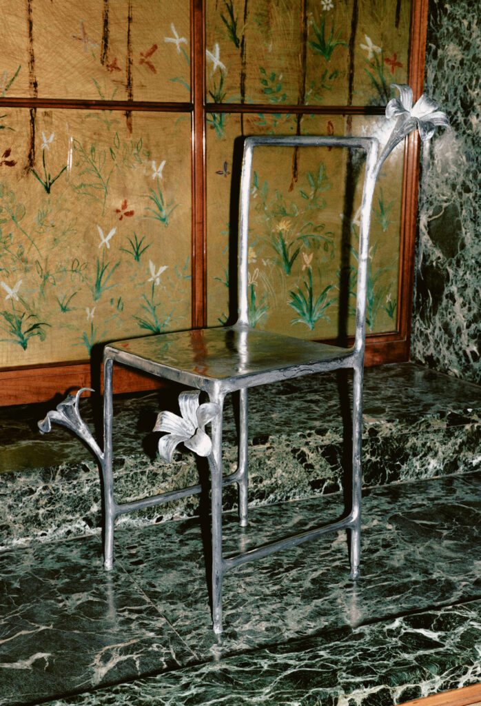 A metallic chair with a minimalist frame and decorative floral accents on the seat and backrest, placed on a green marble surface with a botanical-patterned backdrop.