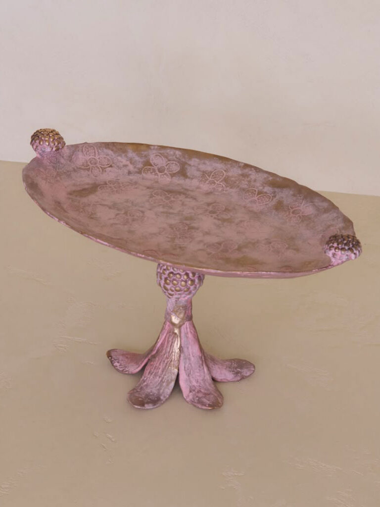 A round, ornate pink metal pedestal tray with embossed floral patterns and decorative knobs on the rim, standing on a three-footed base.