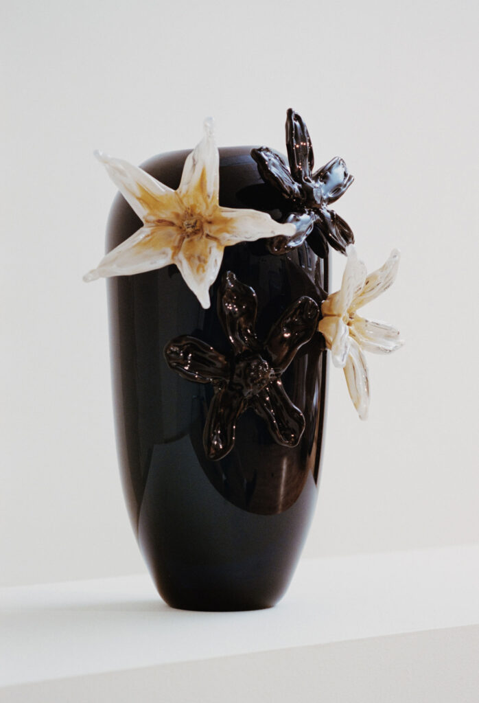 A glossy black vase with translucent glass flowers attached to its surface, displayed against a plain white background.