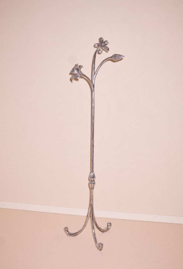 A silver metal coat rack with floral and leaf designs at the top, standing on three curved legs against a plain beige background.