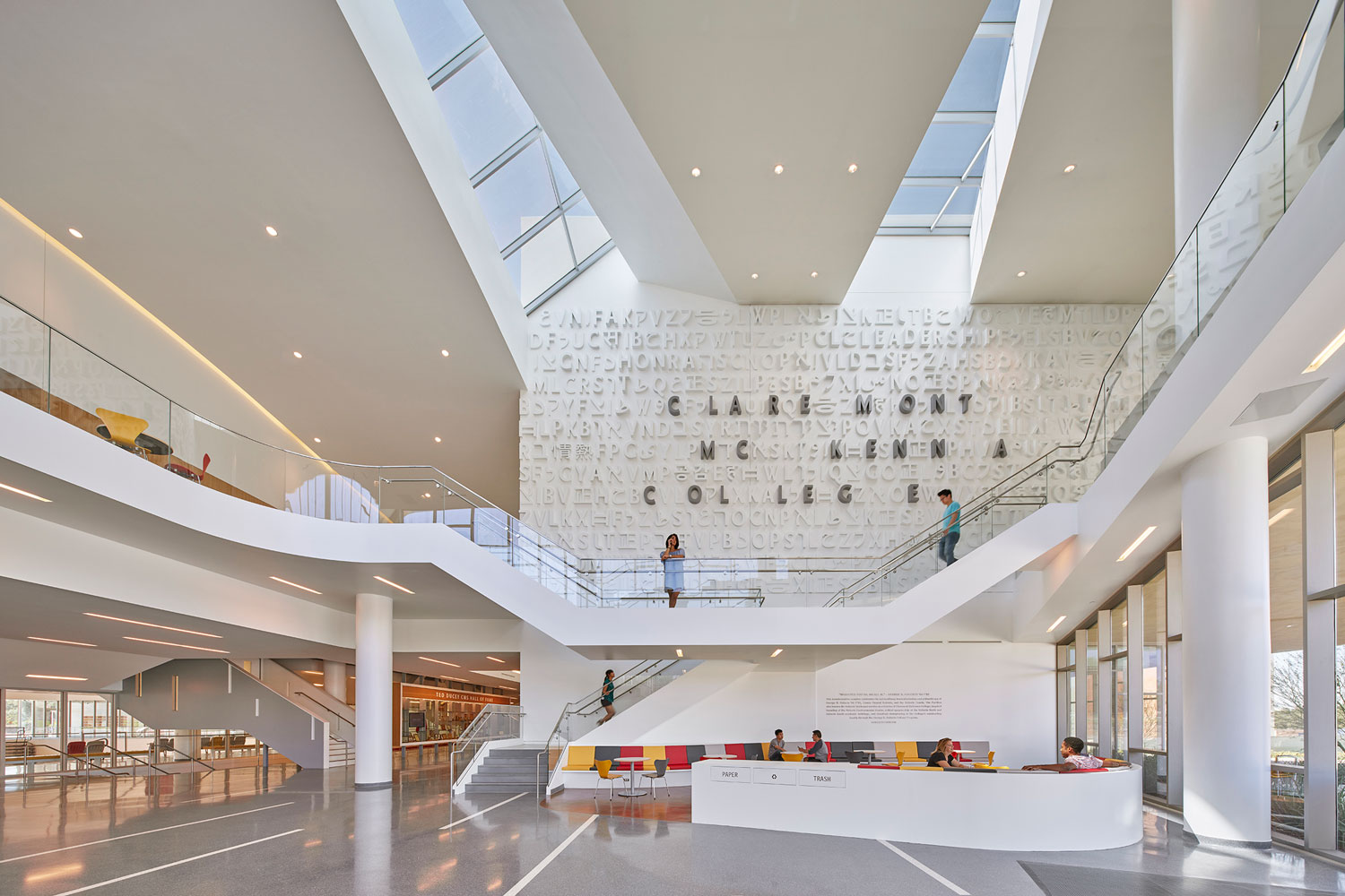 A multi-level, modern library atrium with a large wall feature displaying the letters of “Claremont College” and several people walking on stairs or sitting at a curved reception desk.