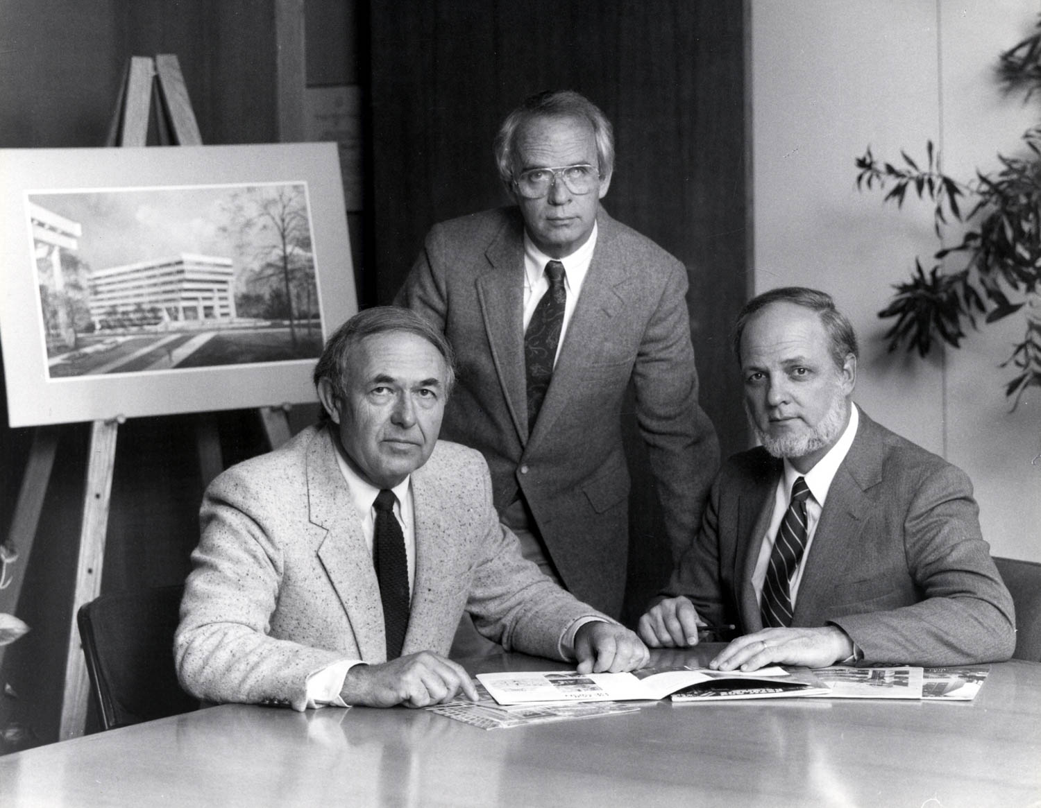 Three men in suits sit and stand around a table with papers; an architectural rendering of a building is displayed on an easel behind them.