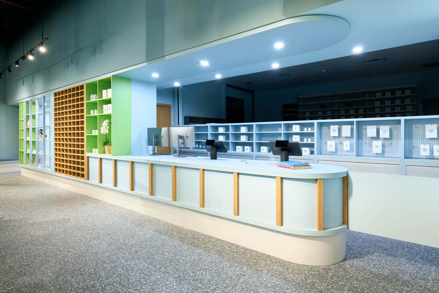 A modern pharmacy counter with two computer stations, shelves of products, and a minimalist green and blue interior design.