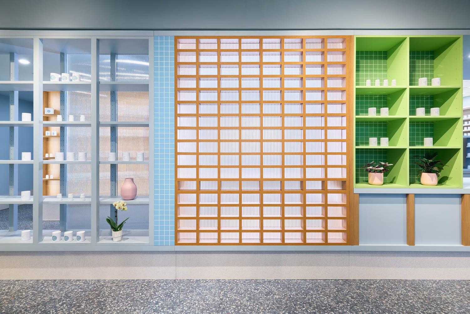 Modern interior with shelves holding small white containers, a pink vase, potted plants, and a wooden grid feature in the center, all against pastel-colored walls and a terrazzo floor.
