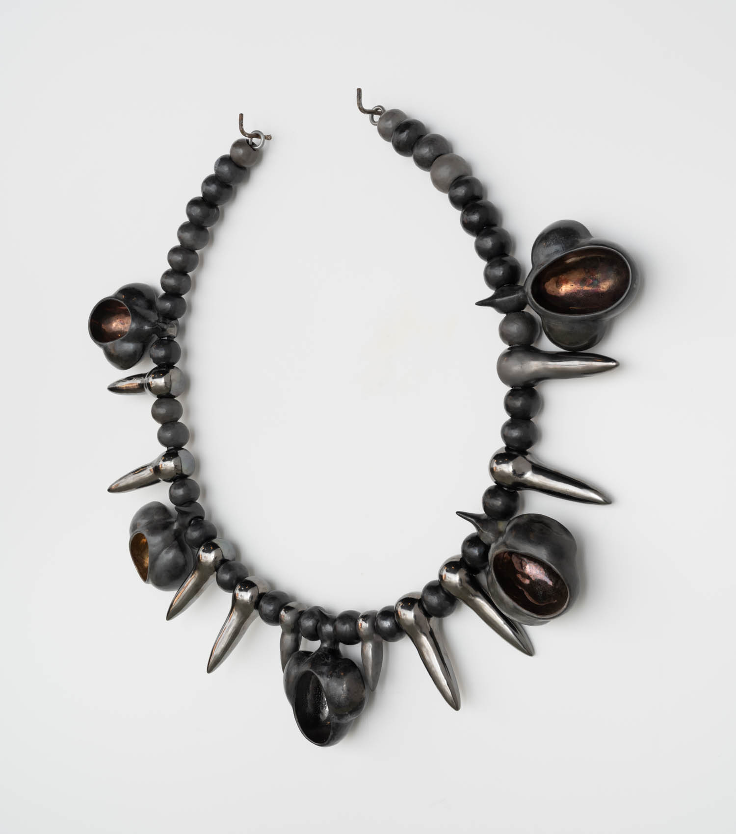 A necklace featuring round, dark beads, metallic spikes, and hollow, shell-like forms arranged symmetrically on a white background.