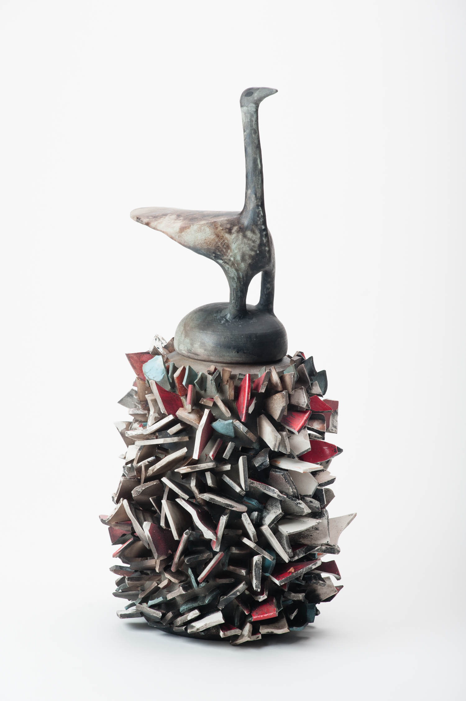 A ceramic sculpture featuring a stylized bird perched atop a base covered with jagged, angular protrusions in shades of gray, red, and white against a white background.