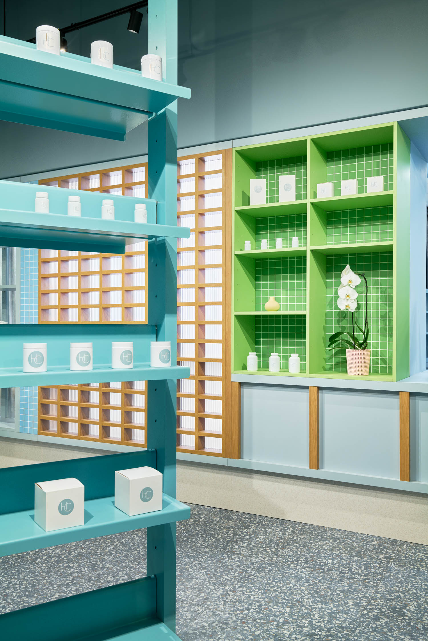 Bright shop interior with teal shelves holding white boxes and a green-tiled counter area with a white orchid in a pink pot, all in a modern, minimalist design.