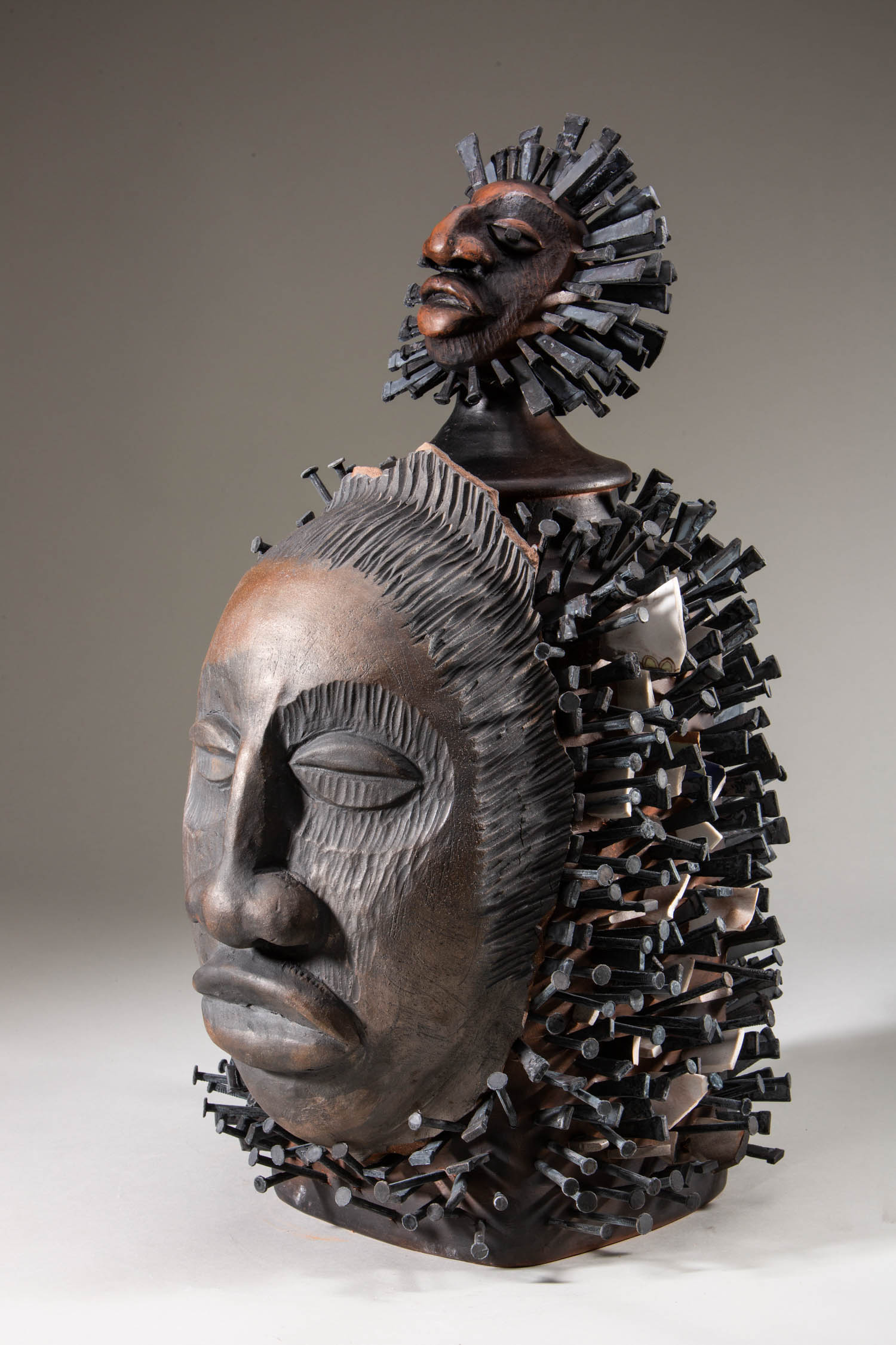 Sculpture featuring two stylized human faces, one above the other, both surrounded by numerous nails protruding outward from the surface.