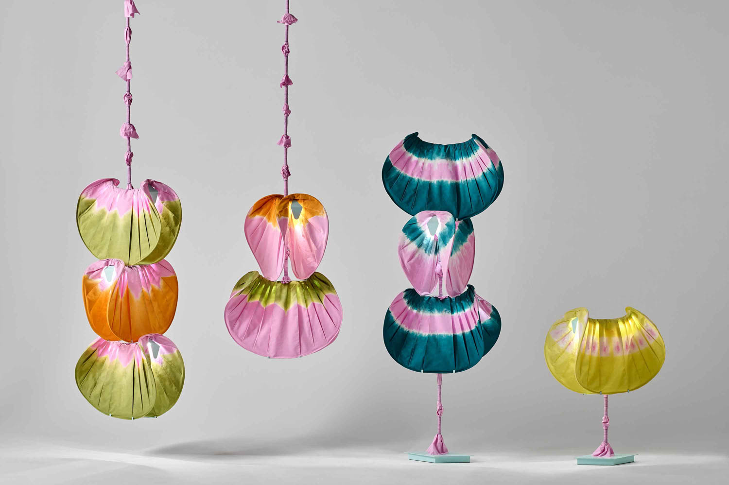 Four colorful, fabric-wrapped sculptural objects with rounded shapes and pastel tones are displayed upright and suspended against a plain gray background.