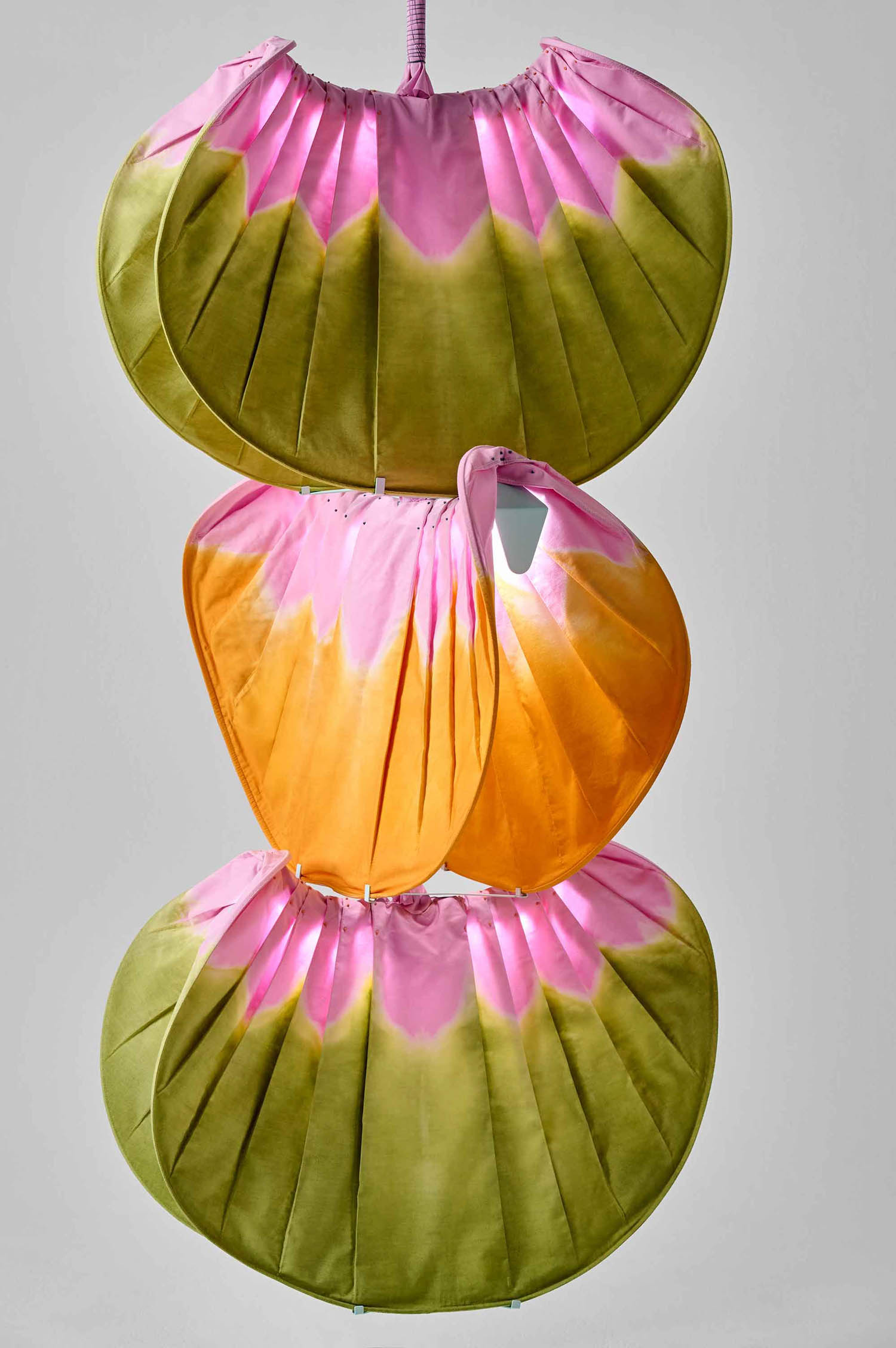 Three pleated, fan-shaped fabric panels in green, pink, and orange are stacked vertically and suspended against a plain background.