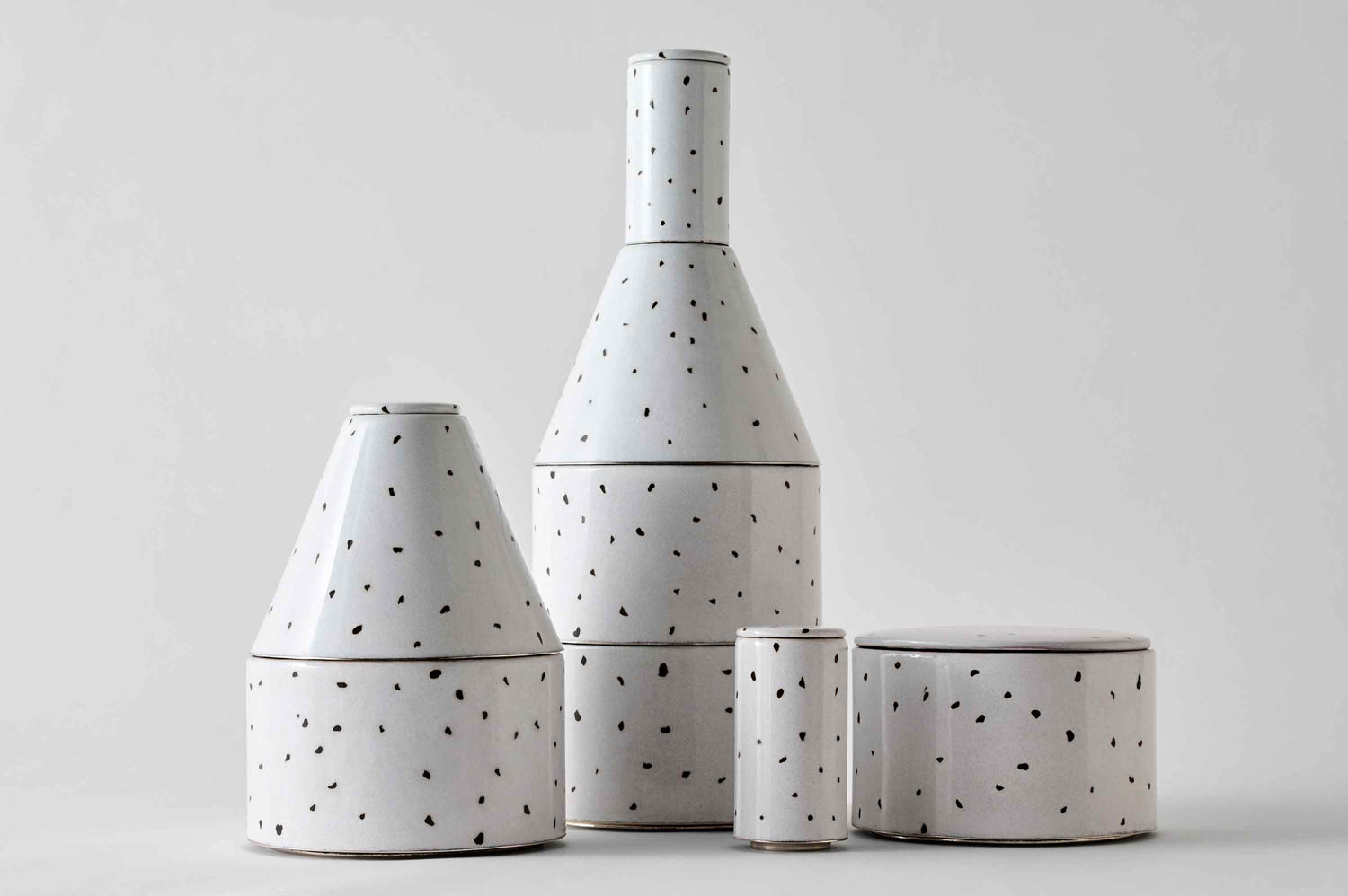 Four white ceramic containers with black speckles, each of different shapes and sizes, are arranged on a plain, light background.