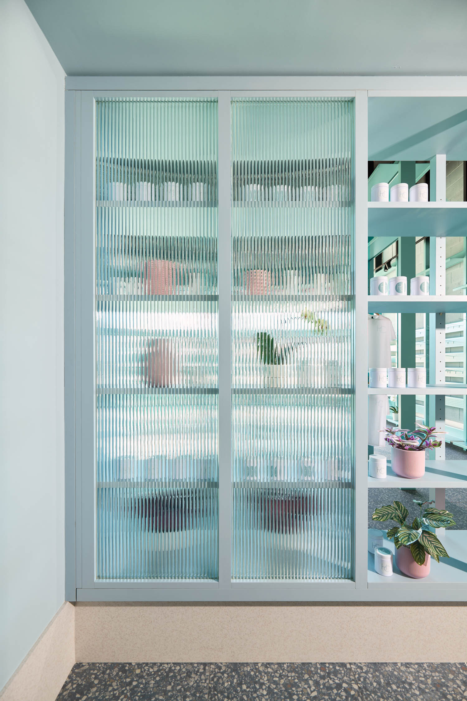 A glass-front cabinet with ribbed panels contains various jars and containers. Nearby shelves display potted plants and cylindrical containers in a modern, light-filled space.