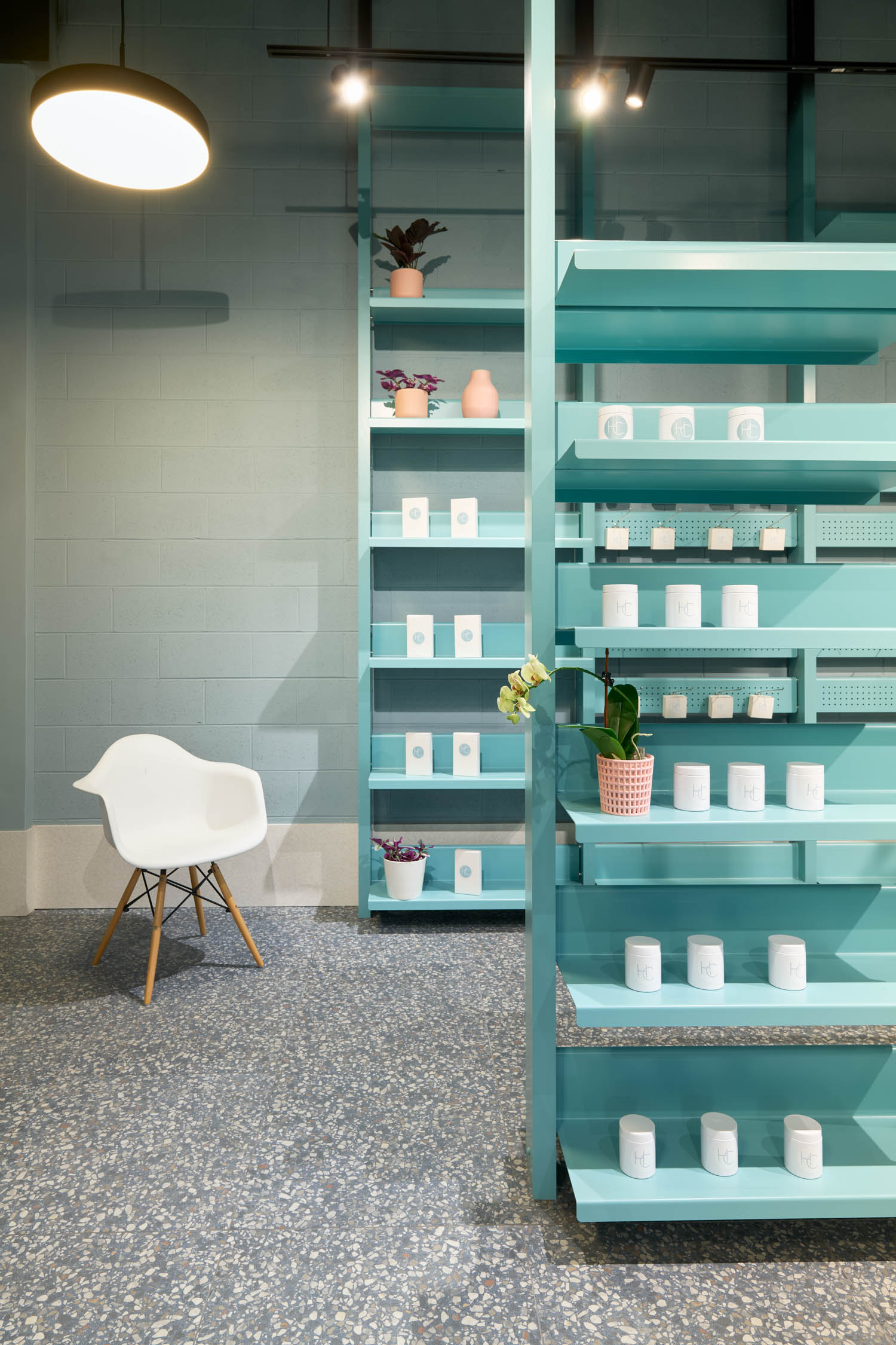 A modern shop interior with turquoise shelves holding white containers and potted plants, next to a white chair on a speckled floor under a large hanging light.