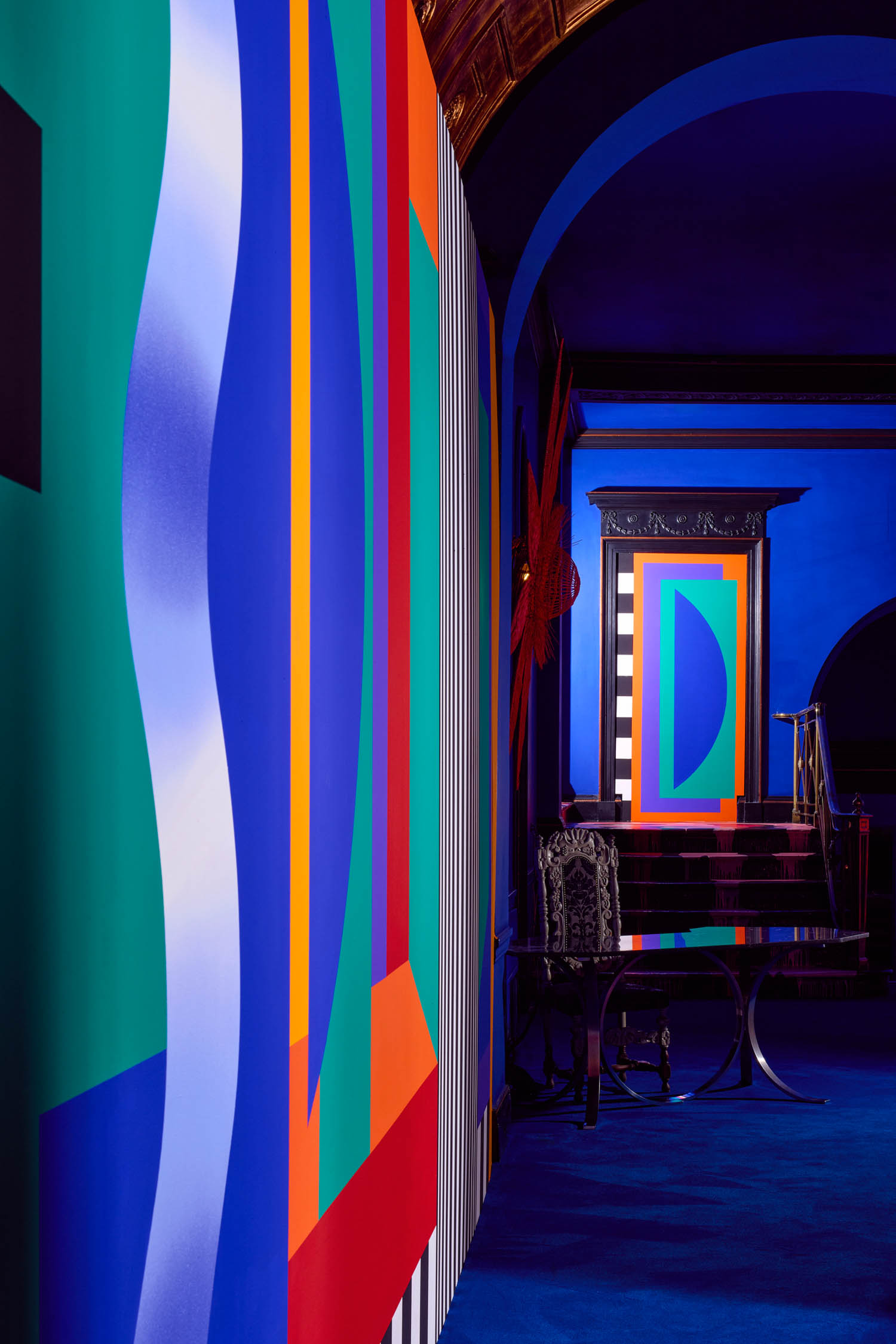 A hallway with bold, geometric wall designs in blue, green, red, orange, and black, leading to a door with a matching colorful pattern and ornate black trims.