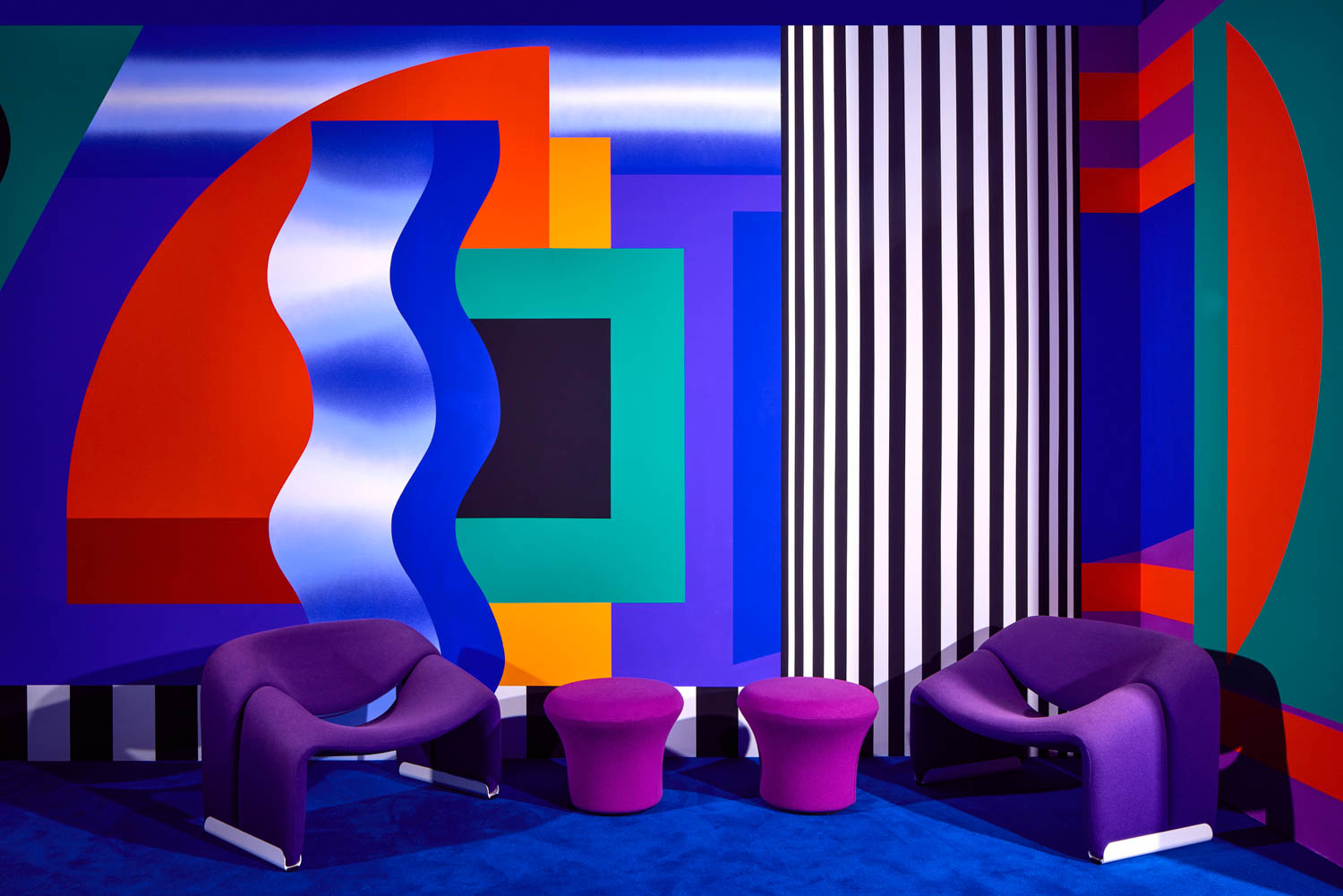 Colorful room with bold geometric wall art, two purple modern chairs, and two matching purple stools on a blue carpet.