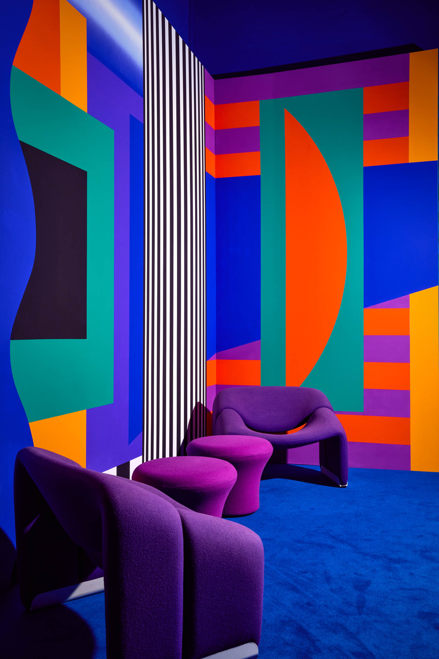 Two purple lounge chairs and a small table sit in a vibrant room with geometric, multicolored wall art and a blue carpet.
