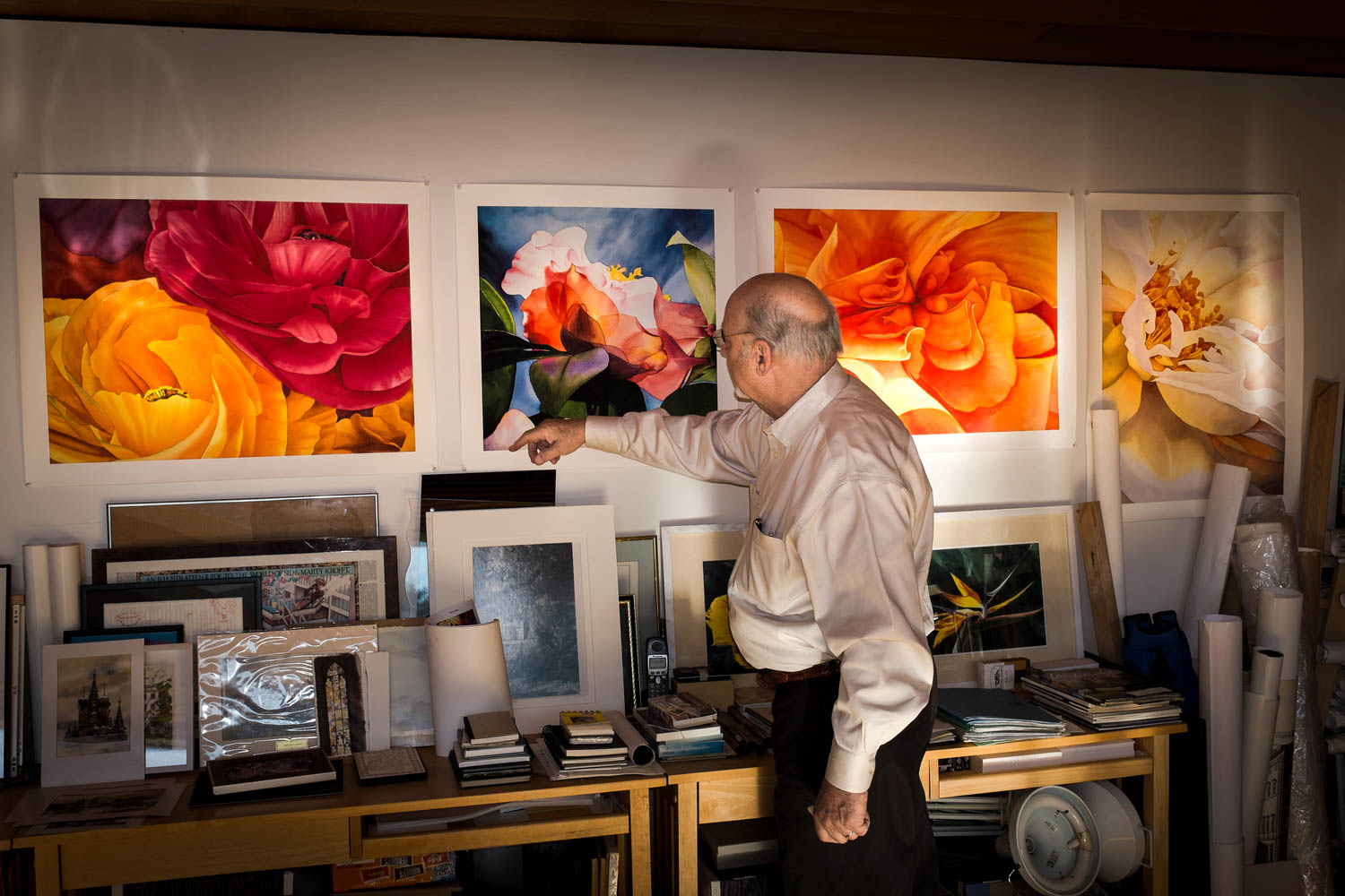 An older man points at a large floral painting on a wall, surrounded by other vibrant flower artworks and art supplies on shelves.