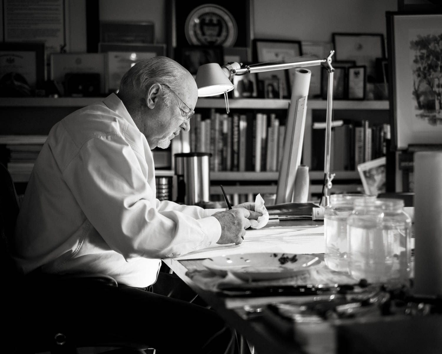 An older man in a white shirt sits at a desk under a lamp, examining papers in a study filled with books, awards, and framed certificates.