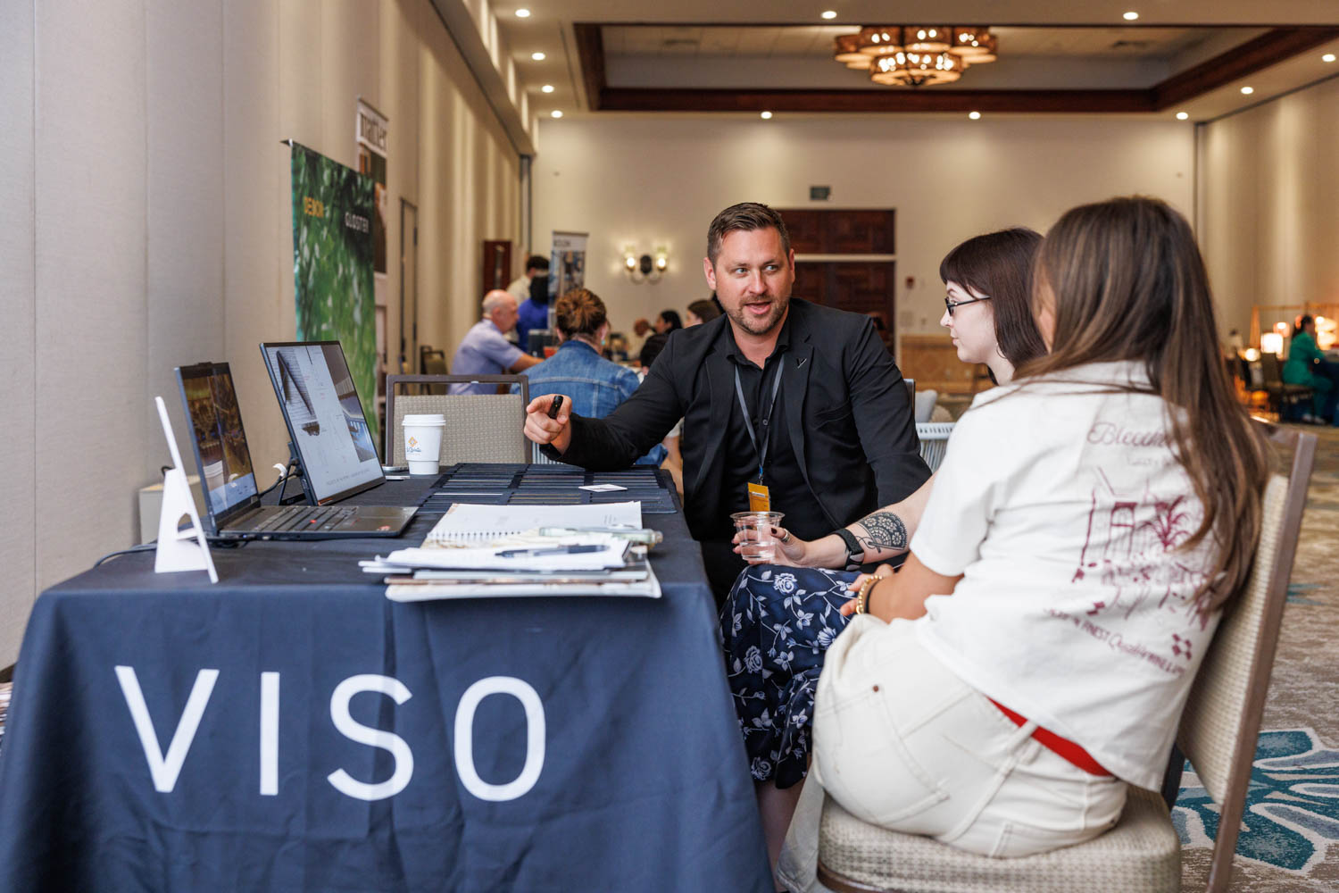 A man talks to two women at a table with laptops and a “VISO” banner in a conference room setting.