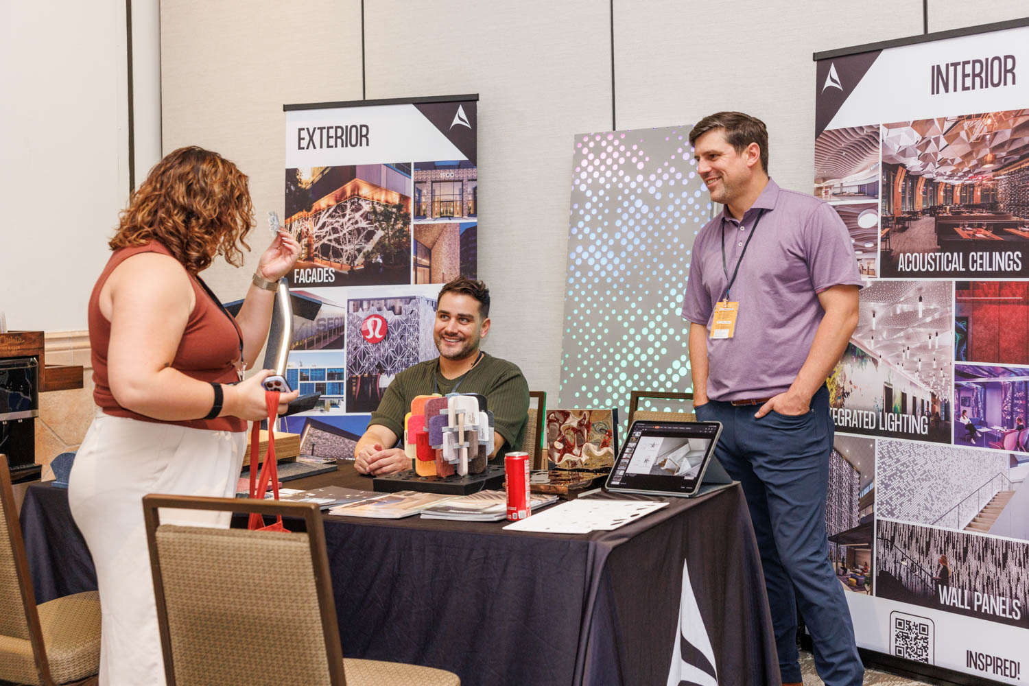 Three people interact at a trade show booth displaying samples and materials for exterior and interior architectural products. Two men stand and sit behind the booth, while a woman speaks with them.