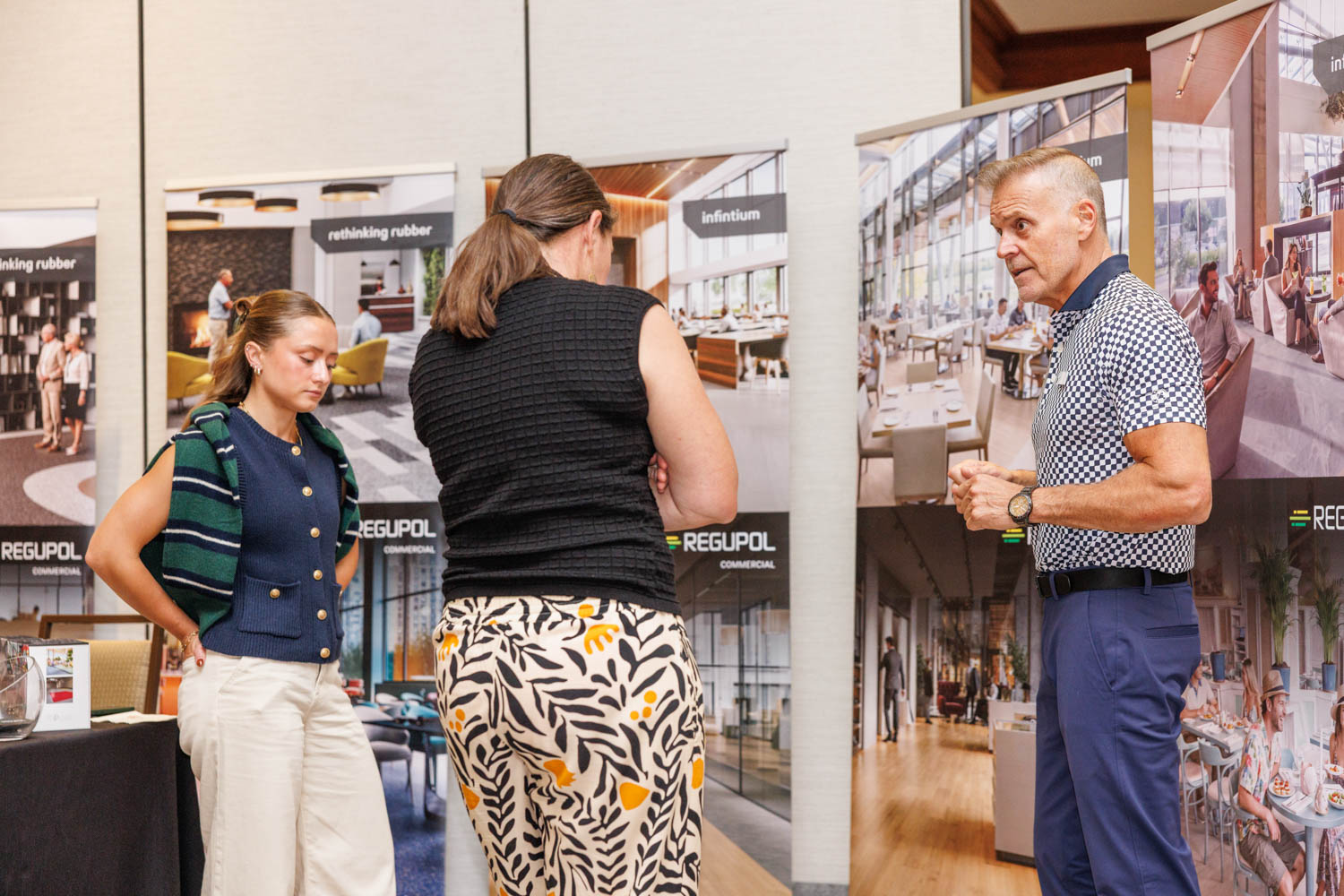 Three people stand and talk in front of display banners at an indoor event or exhibition. The banners feature images of commercial interiors and the word “REGUPOL.”.
