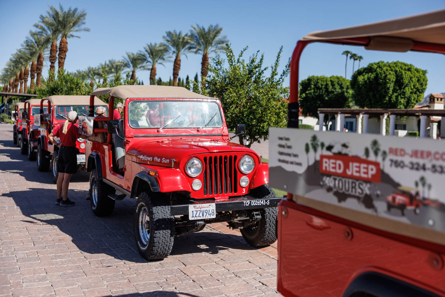 Several red jeeps are parked in a row on a paved area, with a person standing next to one. Palm trees and a building are visible in the background.
