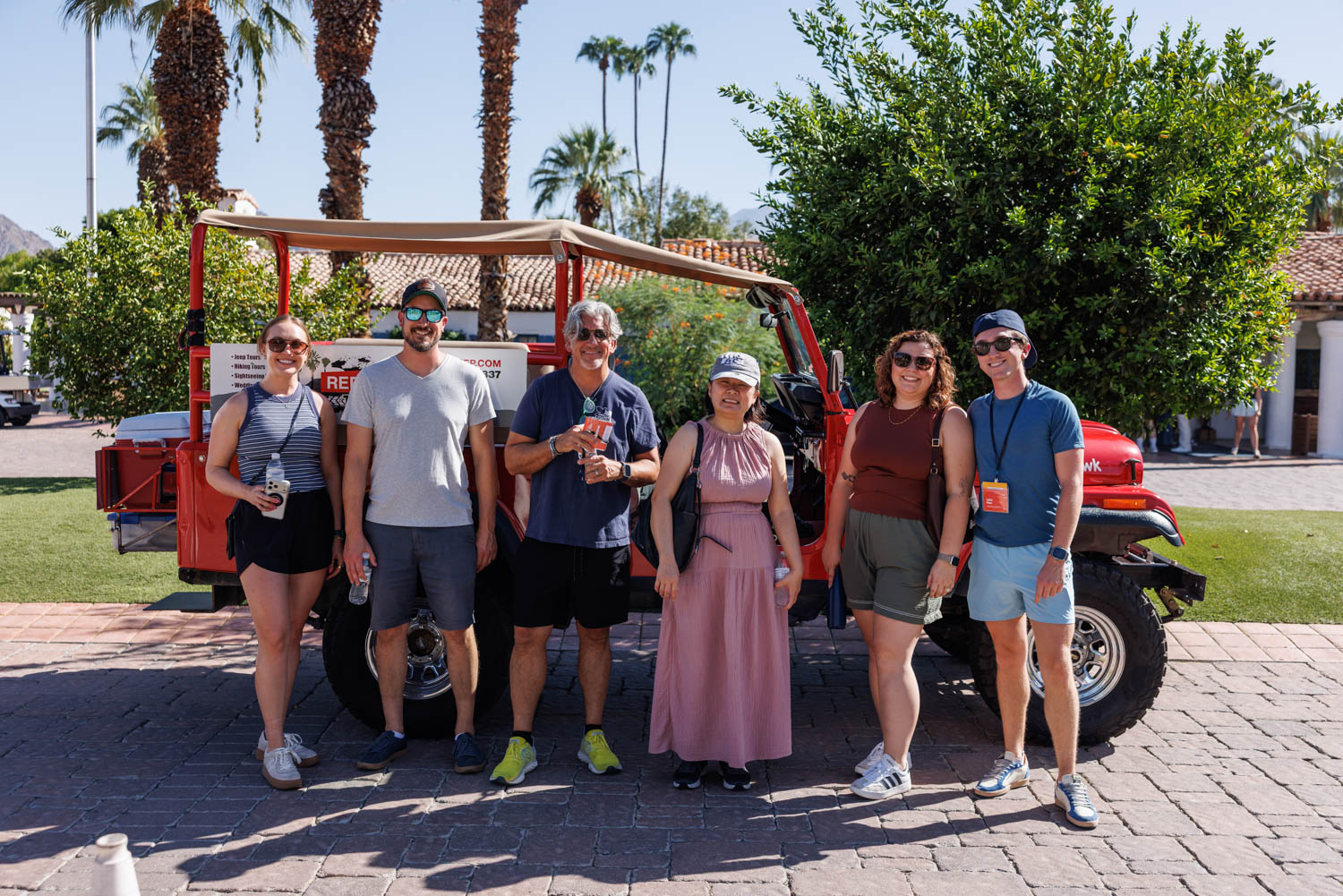 Six people stand in front of a red open-air jeep parked on a paved area, with palm trees, greenery, and buildings in the background on a sunny day.