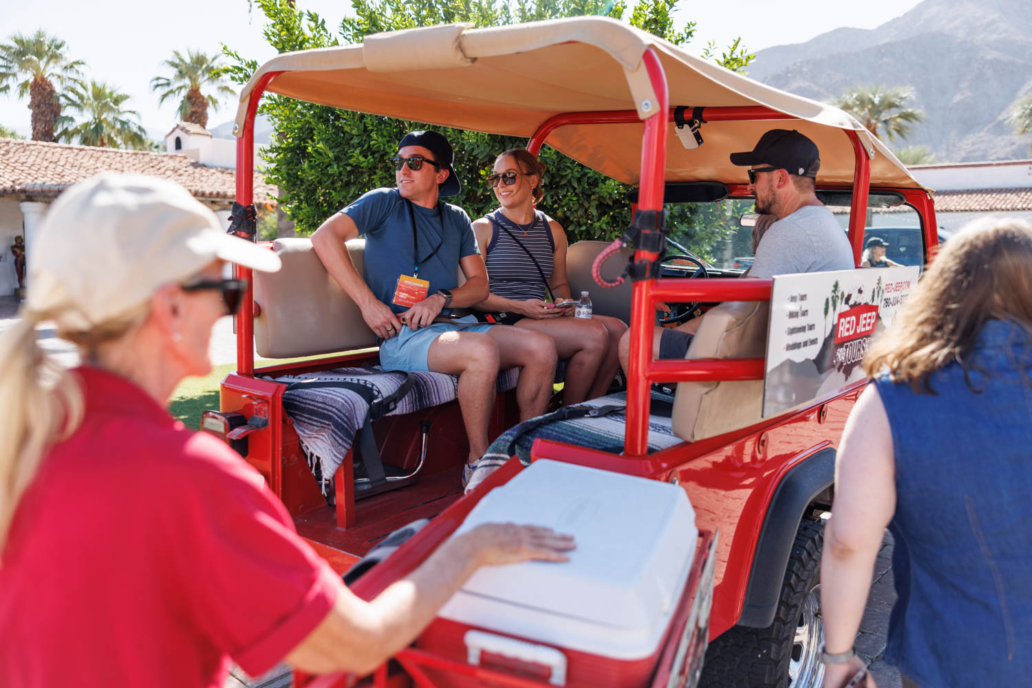 Four people sit in a red open-air tour vehicle while a guide stands nearby outdoors on a sunny day with palm trees in the background.