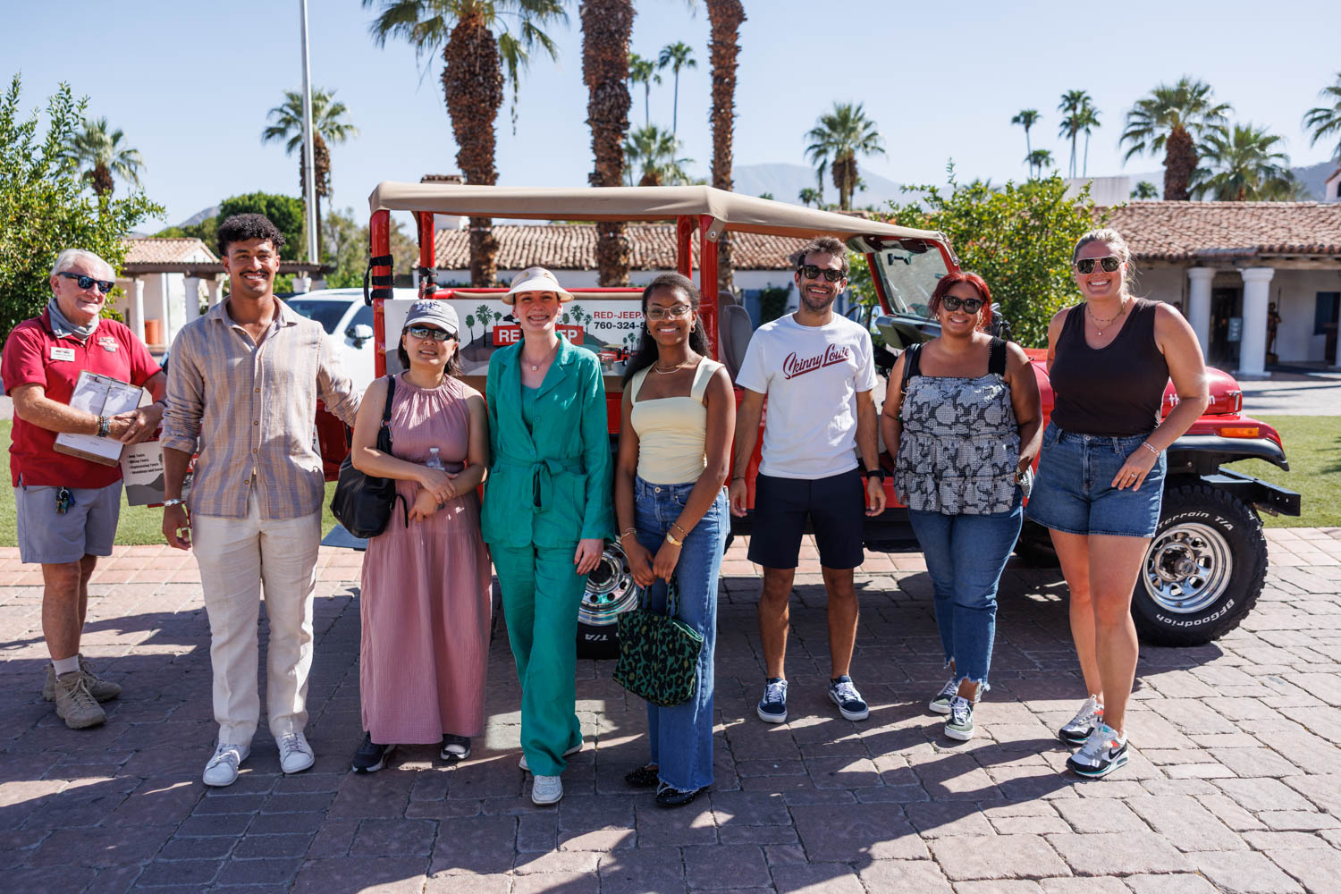 A group of seven people pose in front of a red jeep outdoors on a sunny day, with palm trees and a building in the background.