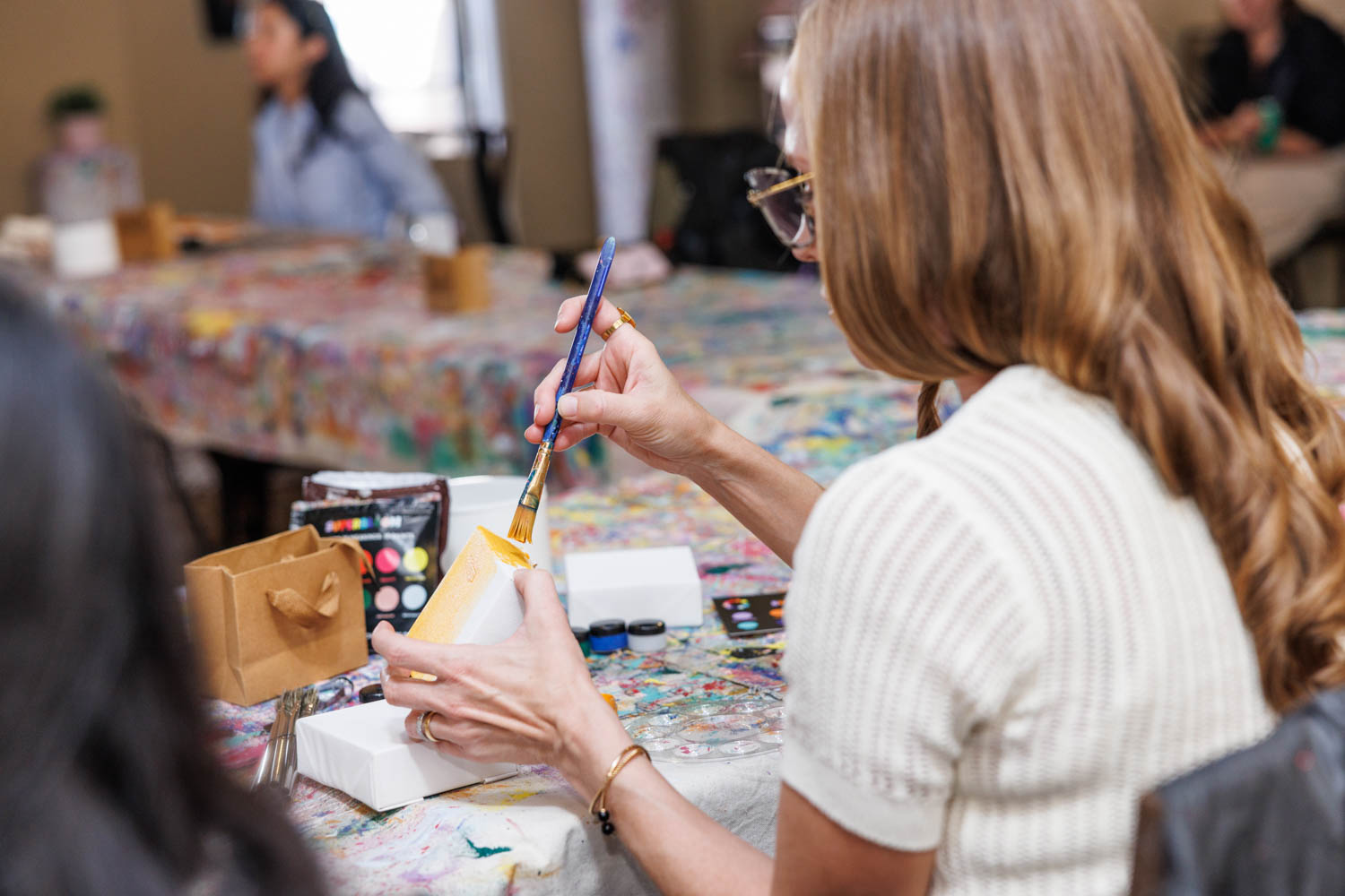 A woman with long hair paints a white ceramic cup at a table covered with art supplies and other people in the background.