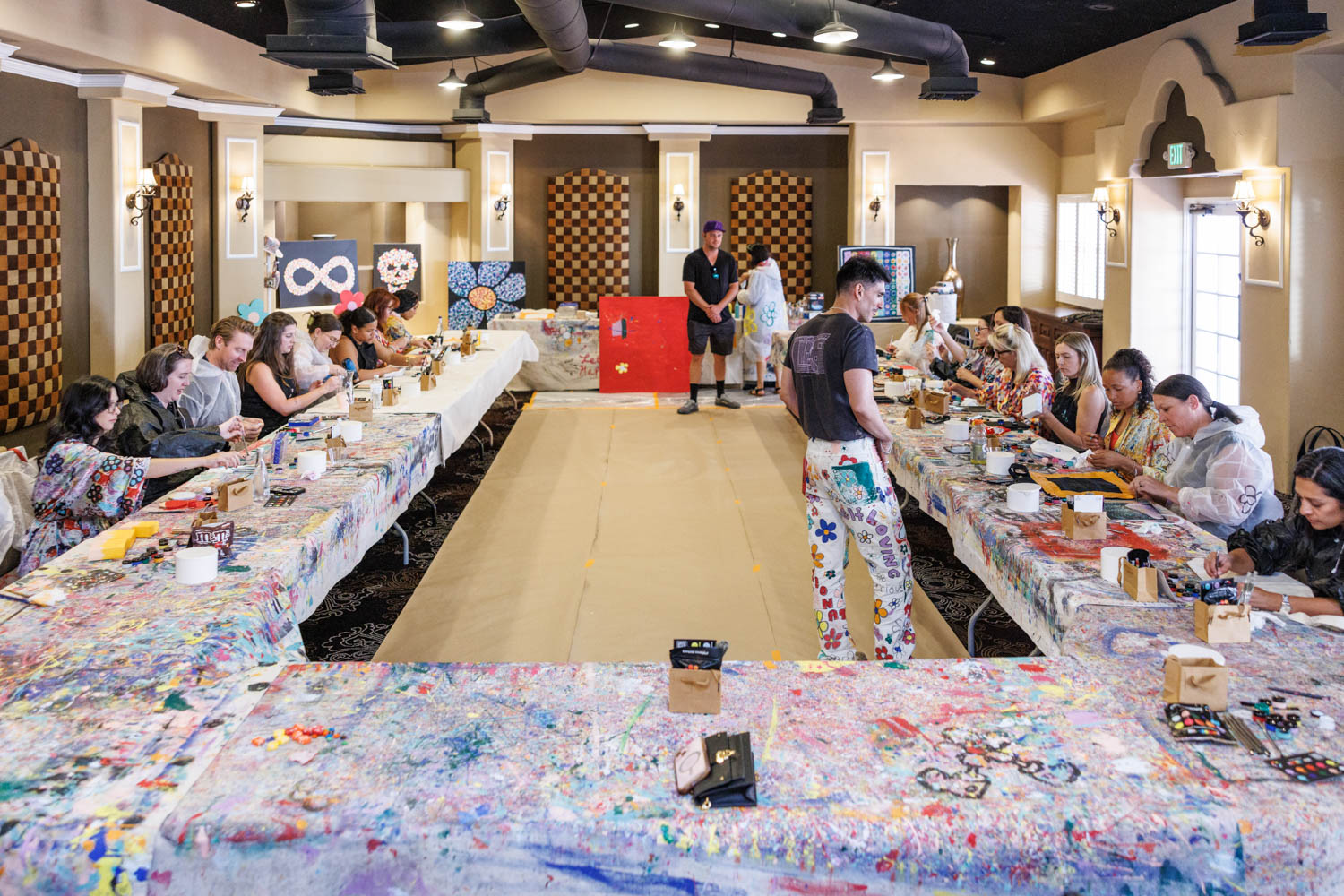 A group of people sit at long tables covered with painted cloths, participating in an indoor art workshop, while two instructors stand at the front of the room.