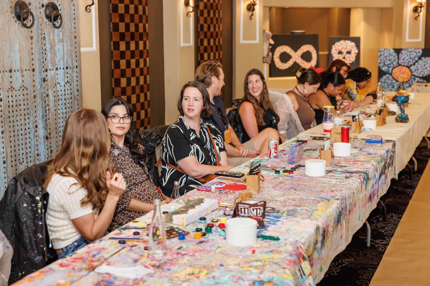 People sit at a long, paint-splattered table with art supplies, talking and working on creative projects in a well-lit indoor space.