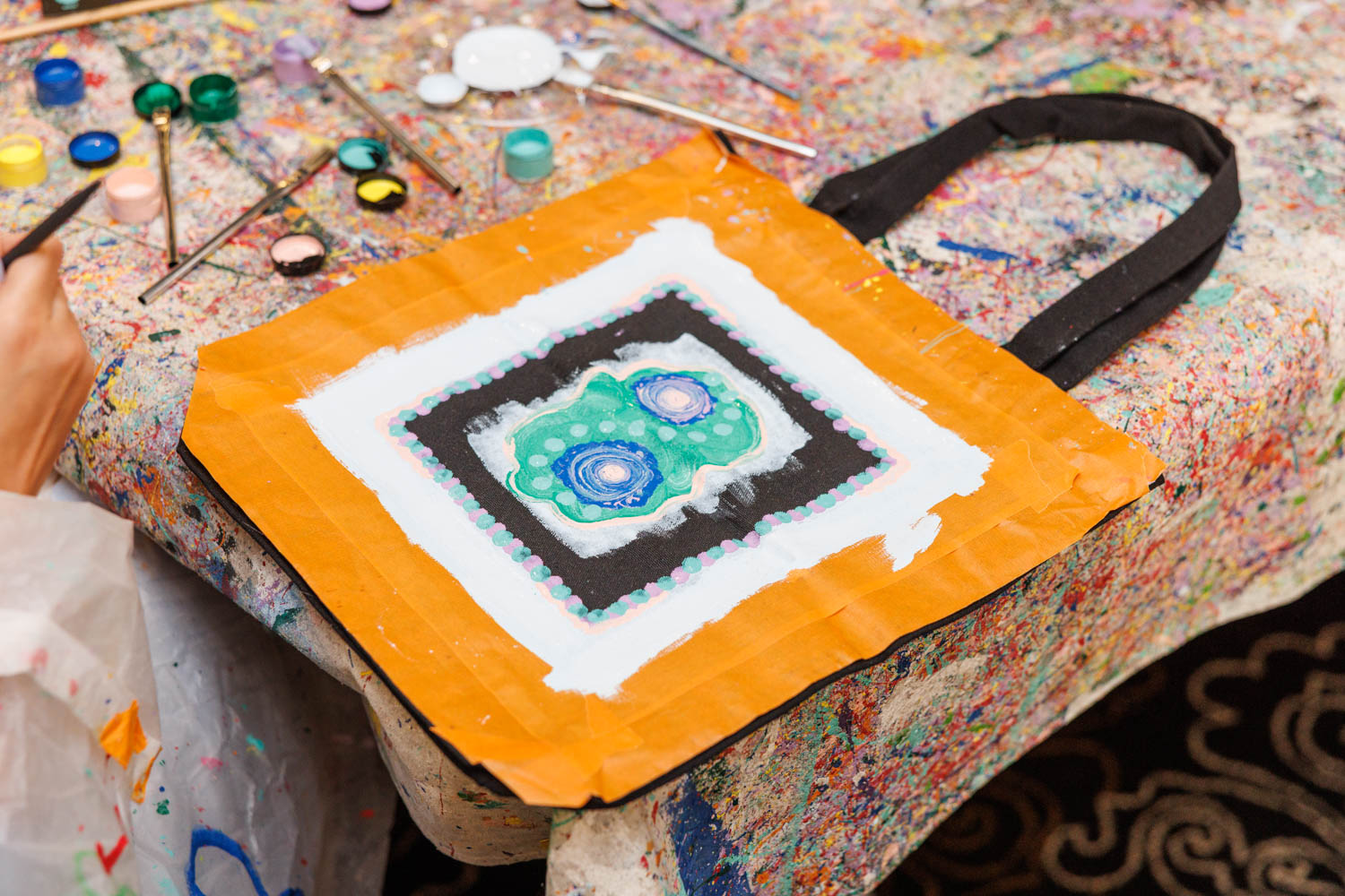 A hand-painted tote bag with an abstract design in blue, green, black, and white on an orange background sits on a paint-stained table with paint pots and brushes nearby.