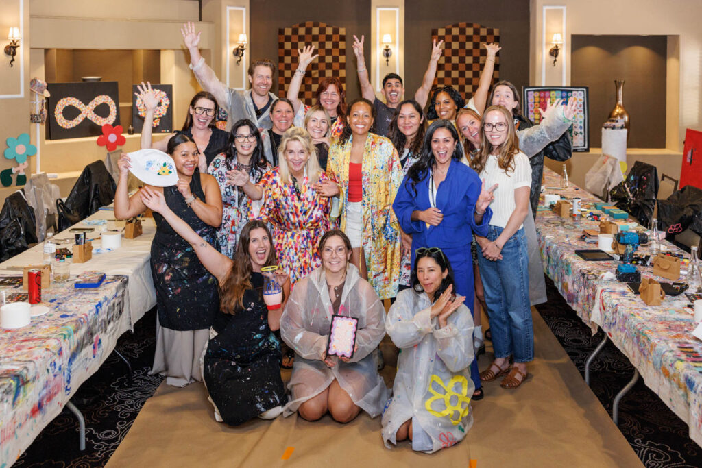 A group of people pose and smile in an art studio, surrounded by tables covered with art supplies, paintings, and decorated items. Some hold up their completed art projects.