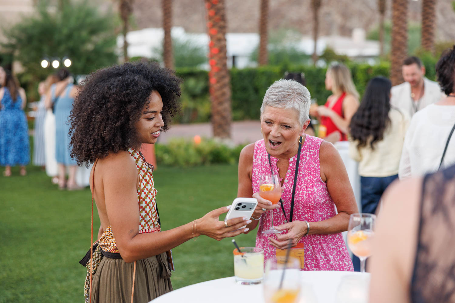 Two women standing outdoors at a social event, looking at a smartphone and holding drinks, with other people mingling in the background.