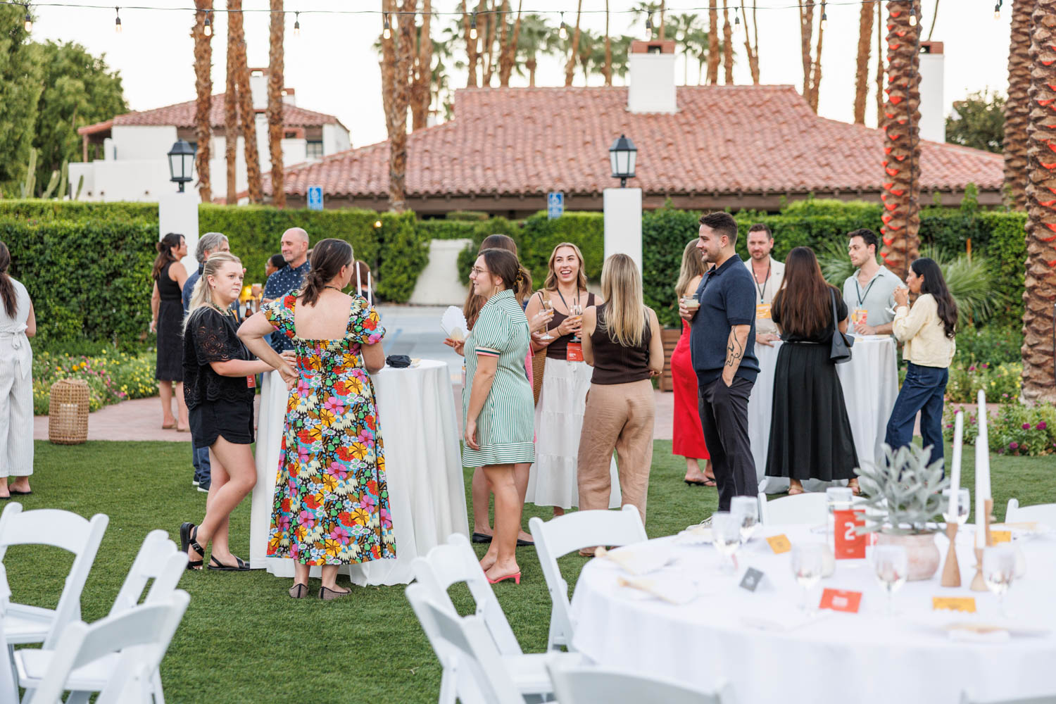 People stand and talk around cocktail tables at an outdoor event on a lawn, with palm trees, string lights, and a building in the background.