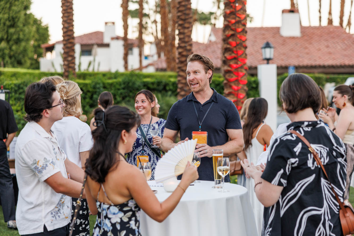 A group of people stand around a cocktail table outdoors, talking and holding drinks, with palm trees and buildings in the background.